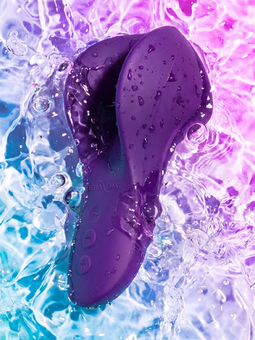 A purple silicone personal massager with a curved, ergonomic shape, shown partially submerged in splashing water with droplets on its surface. The background is a vibrant, split-color water scene transitioning from bright cyan-blue on the left to luminous purple and pink on the right.