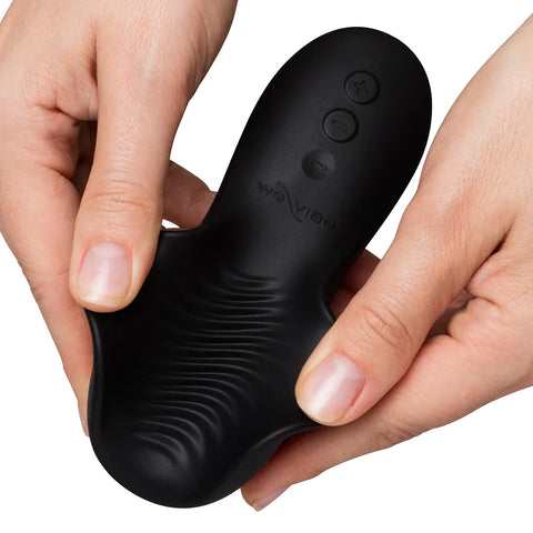 Black personal massager held in two hands against a white background, showing the underside with control buttons and the brand name We-Vibe